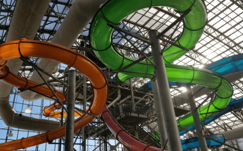 Colorful water slides twist and turn inside a large glass structure, with sunlight streaming through the roof.