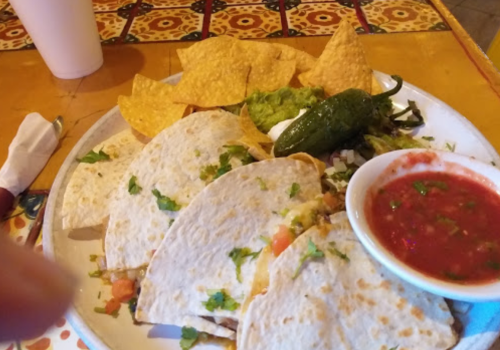 A plate of tacos with salsa, guacamole, chips, and a jalapeño pepper on a colorful table.