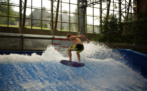 A person surfing on an indoor wave machine, surrounded by splashing water and large windows in the background.