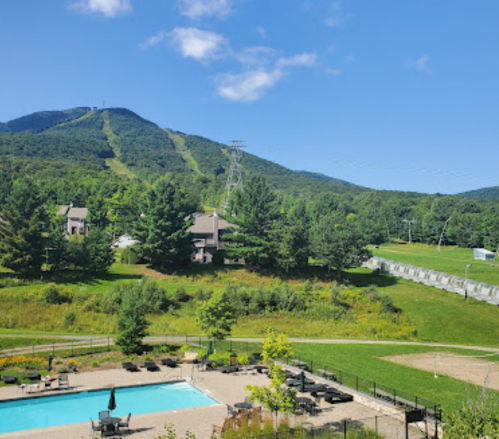 A scenic view of a mountain with ski slopes, a clear blue sky, and a pool area surrounded by greenery.