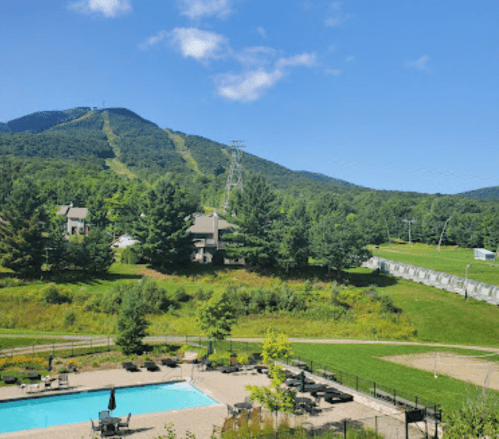 A scenic view of a mountain with ski slopes, a clear blue sky, and a pool area surrounded by greenery.