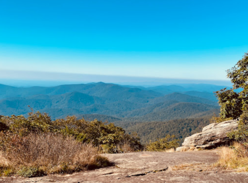A panoramic view of rolling mountains under a clear blue sky, with rocky terrain in the foreground.