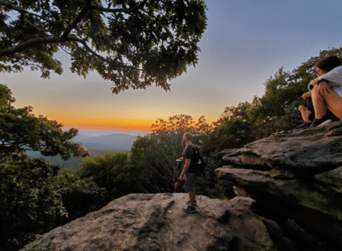 A person stands on a rock ledge, gazing at a colorful sunset over a mountainous landscape, with another person seated nearby.