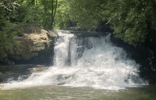 A serene waterfall cascades over rocks, surrounded by lush green trees and a tranquil pool below.
