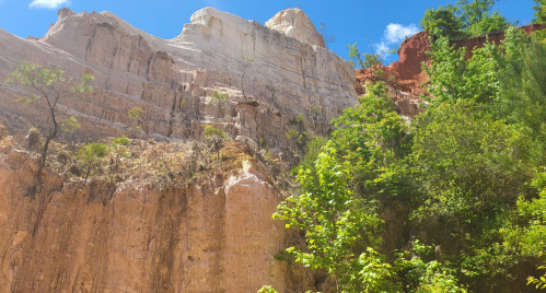 A scenic view of tall, layered cliffs with white and red rock formations, surrounded by lush green vegetation and blue sky.