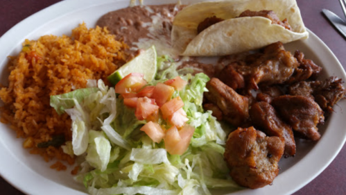 A plate of Mexican food featuring rice, refried beans, tacos, grilled meat, lettuce, and diced tomatoes.