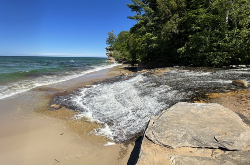 A serene beach scene with gentle waves lapping at the shore, surrounded by lush greenery and rocky formations.