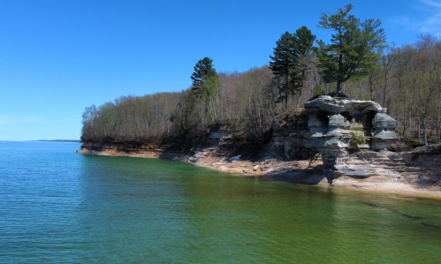 A serene lakeside view featuring rocky cliffs, green water, and trees under a clear blue sky.