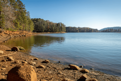 A serene lakeside view with calm water, rocky shore, and trees reflecting autumn colors under a clear blue sky.