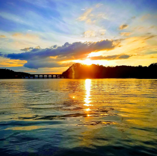 Sunset over a calm lake, with vibrant colors reflecting on the water and a bridge in the distance.
