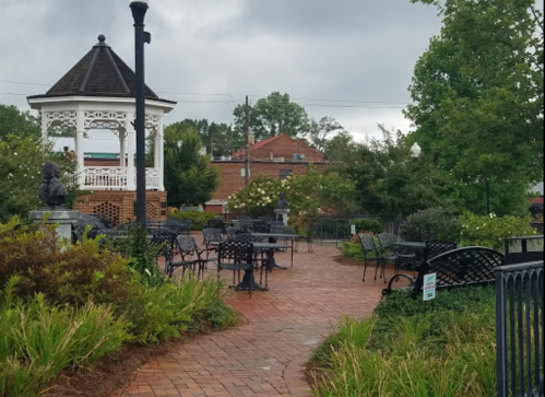 A park scene featuring a gazebo, brick pathway, and benches surrounded by greenery under a cloudy sky.