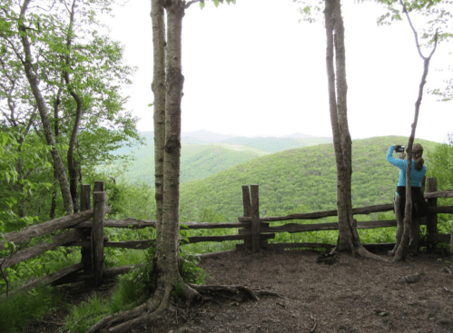 A person stands by a wooden fence, overlooking lush green mountains under a cloudy sky. Trees frame the view.