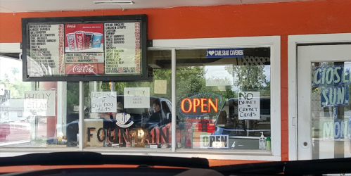 A drive-thru window with a menu board, "OPEN" sign, and various handwritten signs on the glass.