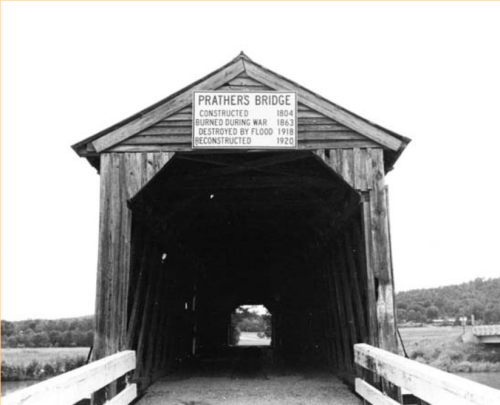 Black and white image of Prathers Bridge, a covered bridge with a sign detailing its history and reconstruction dates.