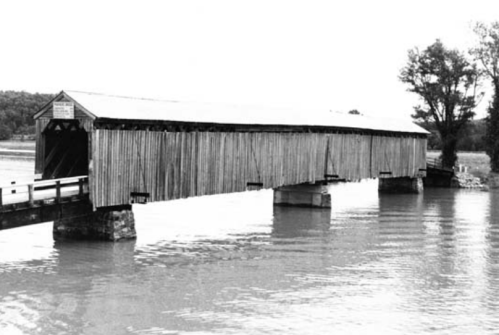 A historic covered bridge spans a river, surrounded by trees, captured in black and white.