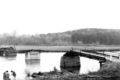 Black and white image of a partially collapsed bridge over a river, with two people examining the structure nearby.