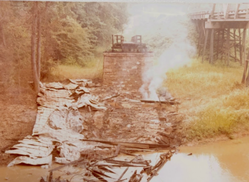 A weathered stone structure with smoke rising, surrounded by debris and overgrown vegetation near a body of water.
