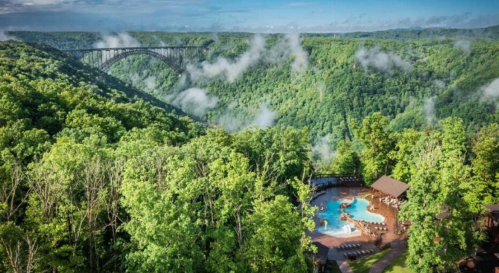 Aerial view of a lush green landscape with a pool area and a bridge spanning a valley under a cloudy sky.