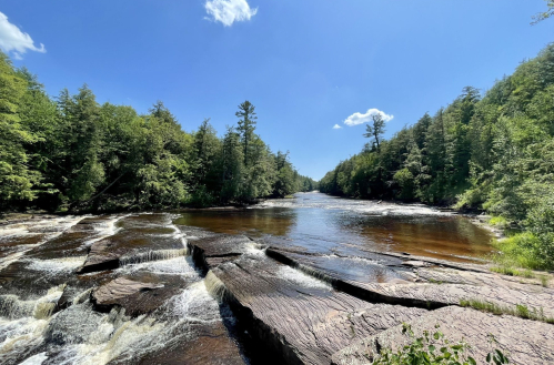 A serene river flows through a lush green landscape under a clear blue sky, with rocky outcrops and gentle rapids.