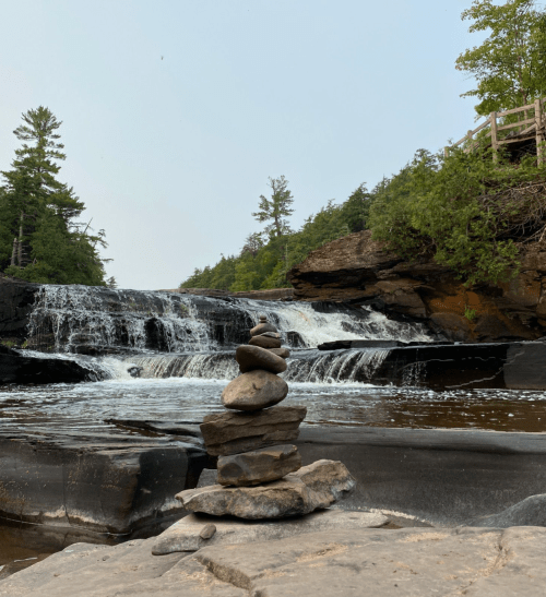 A serene waterfall surrounded by trees, with a stack of smooth stones in the foreground on a rocky surface.