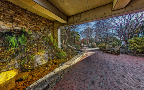 A serene garden view featuring a stone wall, water feature, and brick pathway leading to a peaceful outdoor seating area.