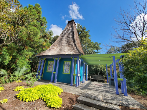A colorful, whimsical building with a pointed roof surrounded by greenery and vibrant plants under a clear blue sky.