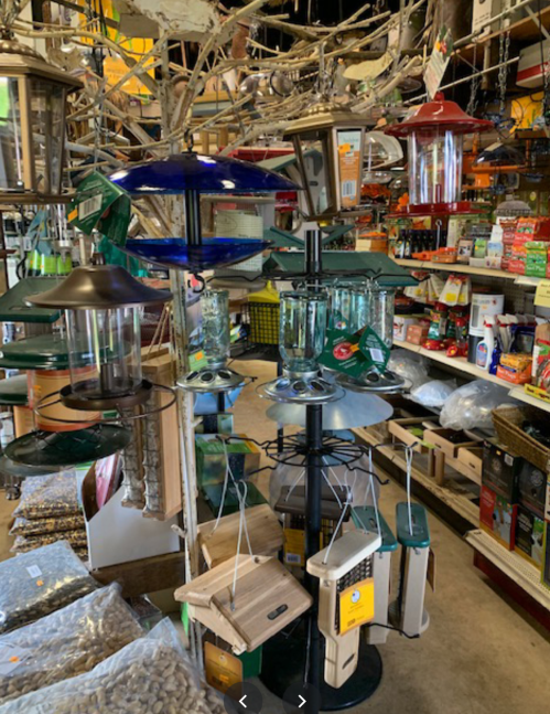 A variety of bird feeders and supplies displayed in a store, surrounded by bags of birdseed and other products.