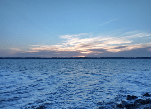 A serene view of a calm ocean at sunset, with soft clouds and a gradient sky reflecting on the water.