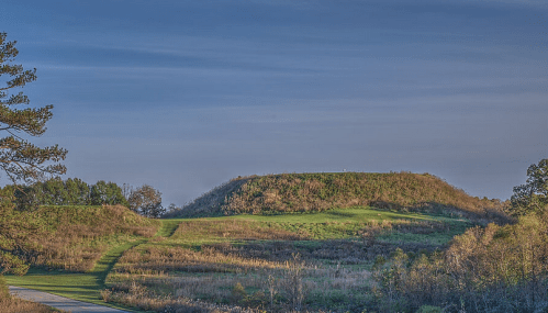 A grassy hill with a gentle slope, surrounded by trees and open fields under a clear blue sky.
