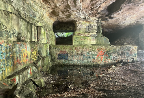 Graffiti-covered rock walls inside a cave, with reflections in shallow water on the ground. Natural light filters in.