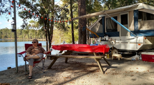A person relaxes in a chair by a lakeside campsite with a picnic table and a camper in the background.