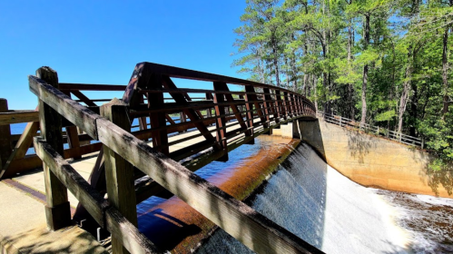A wooden bridge over a dam, with water cascading down and surrounded by lush green trees under a clear blue sky.