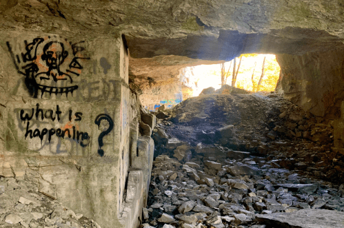 A rocky cave entrance with graffiti on the wall, featuring a figure and the text "what is happenin?" illuminated by sunlight.