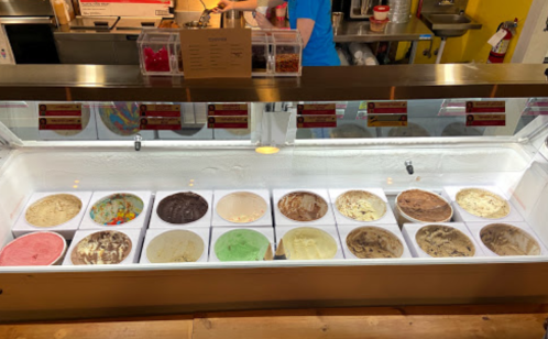 A display of various ice cream flavors in bowls, with a person serving in the background.