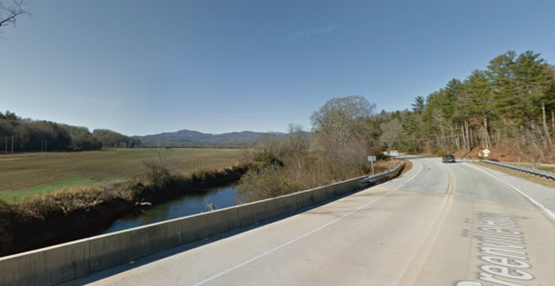 A scenic view of a road alongside a river, with mountains and trees in the background under a clear blue sky.