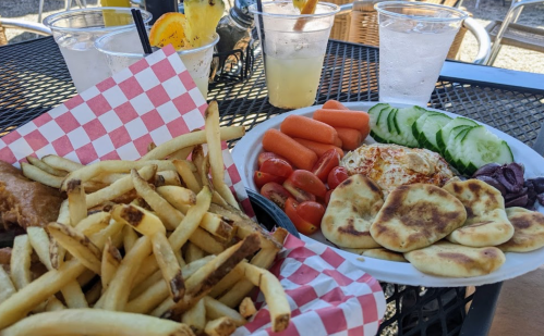 A plate of fries, a colorful vegetable platter with cucumbers, tomatoes, carrots, and pita, alongside drinks on a table.