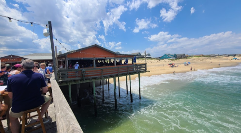 A seaside restaurant on a pier with diners, overlooking a sandy beach and ocean under a partly cloudy sky.