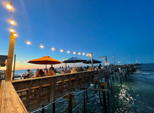 A wooden pier at sunset with diners under umbrellas, string lights illuminating the scene over the ocean.
