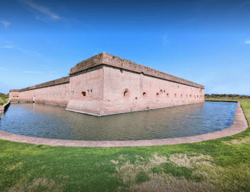 A large, historic brick fort surrounded by water, set against a clear blue sky and green grass.