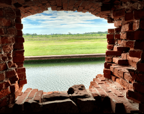 View through a brick archway overlooking a grassy landscape and calm water.