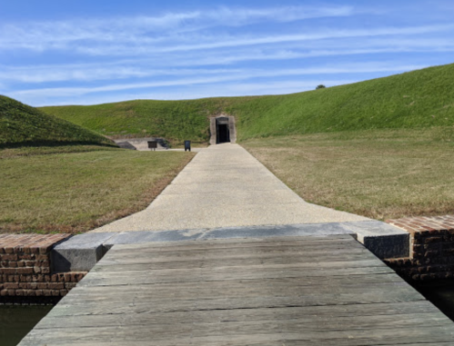 A stone pathway leads to a grassy area with a historic fort entrance under a blue sky.
