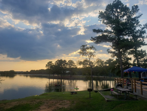A serene lake at sunset, surrounded by trees and a picnic area, with clouds reflecting in the water.