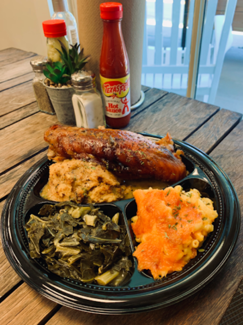 A black plate with sausage, collard greens, macaroni and cheese, and cornbread, with hot sauce in the background.