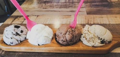 Four scoops of ice cream in a wooden tray: cookies and cream, vanilla, chocolate, and a light-colored flavor, with pink spoons.