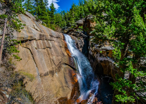 A waterfall cascading down rocky terrain, surrounded by lush green trees and a clear blue sky.