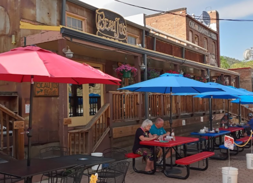A cozy outdoor dining area with colorful umbrellas, featuring two people at a table in front of a rustic restaurant.