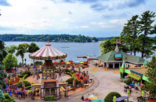 A colorful carousel surrounded by greenery, with a lake and cloudy sky in the background.