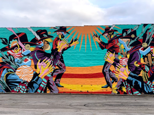 Colorful mural depicting masked figures in a dynamic dance, surrounded by vibrant patterns and a sunburst backdrop.