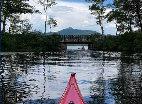 A red kayak on calm water, with a bridge and mountains in the background under a blue sky with clouds.