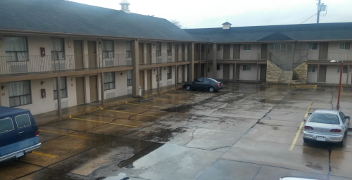 A view of a motel parking lot with wet pavement, featuring two parked cars and a two-story building in the background.